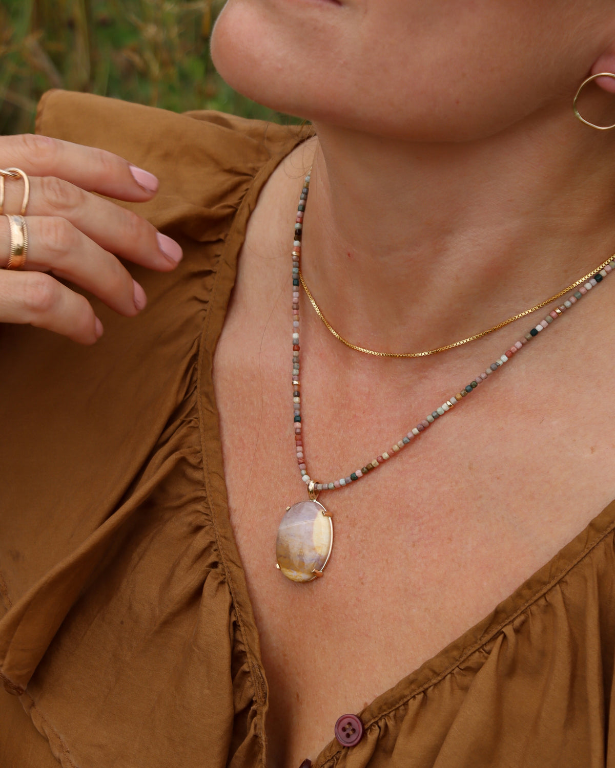 Model wearing a multicolored gemstone beaded necklace with pink, green, cream, and earthy-toned beads, layered with a gold chain and oval gemstone pendant.