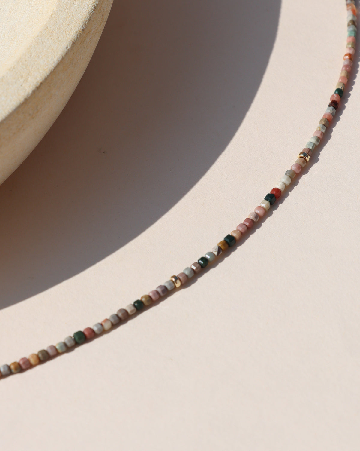 Close-up of a multicolored gemstone necklace featuring tiny faceted square beads in pink, green, cream, brown, and rust tones with gold accents.