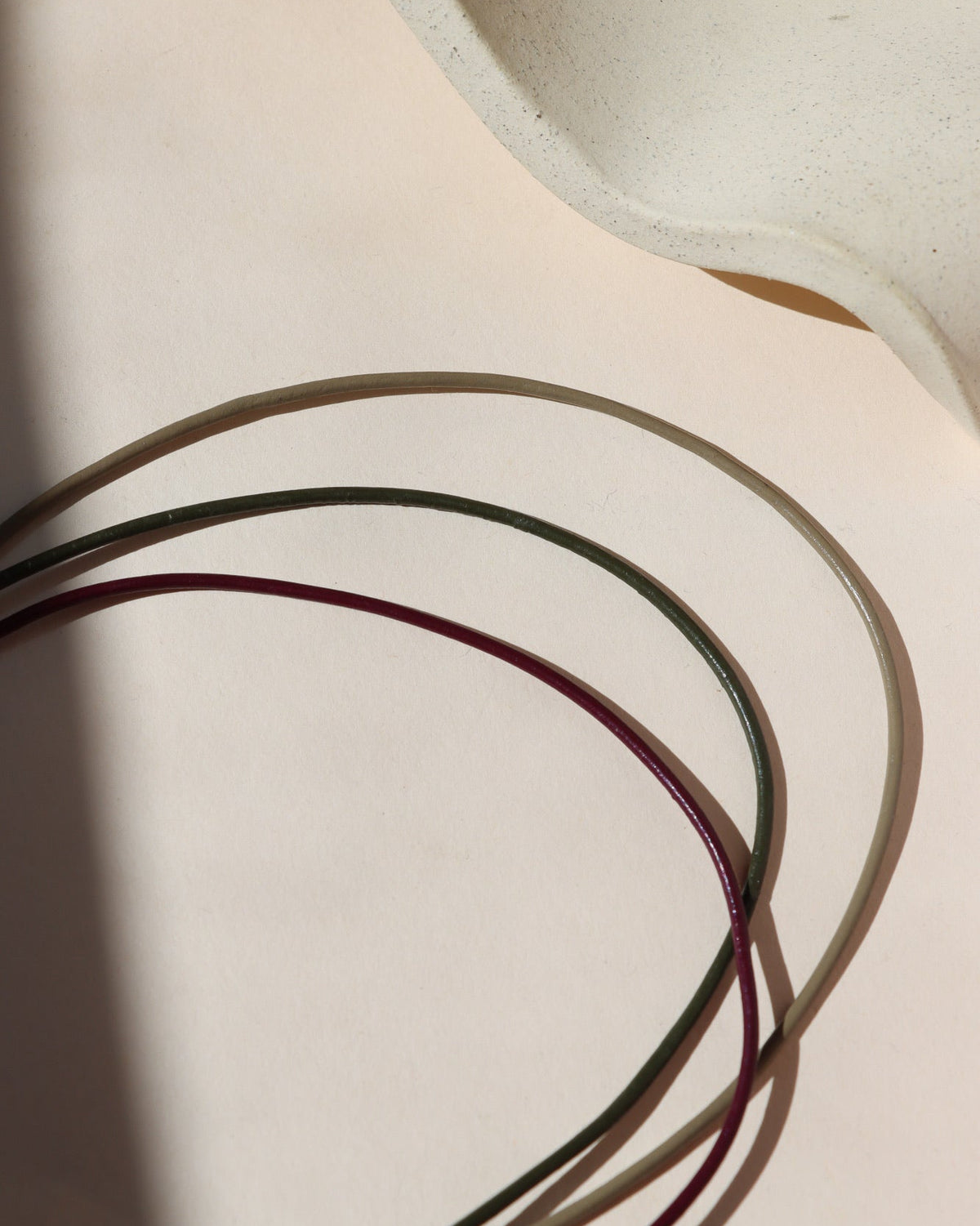 Three thin leather cord necklaces in sand, olive, and plum, arranged in overlapping curves on a cream background in warm sunlight.