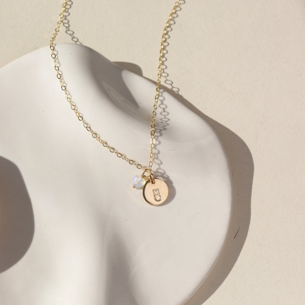 A 14k gold fill simple chain with a stamped disc with a small bunny, paired with a small opal wire wrapped gemstone. Necklace is displayed on a cream backdrop. 