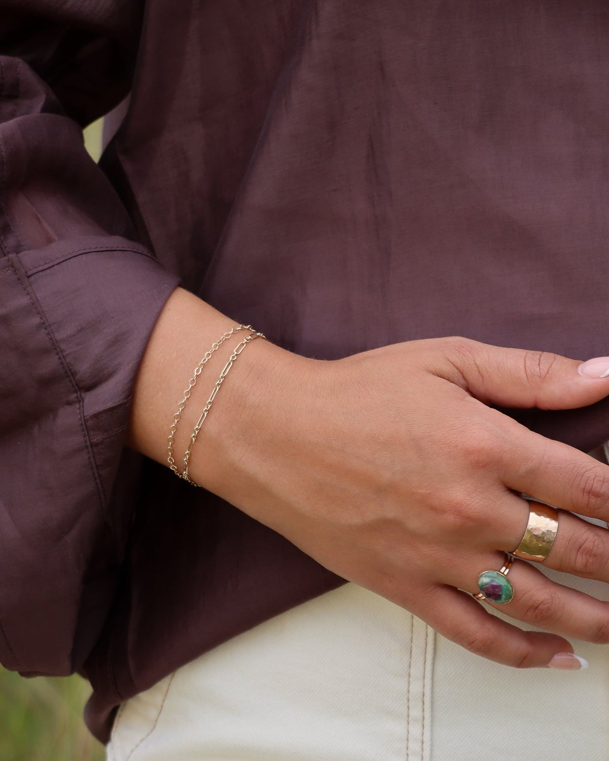 Close-up of a hand wearing two delicate gold chain bracelets and layered rings, including a green and purple gemstone ring.