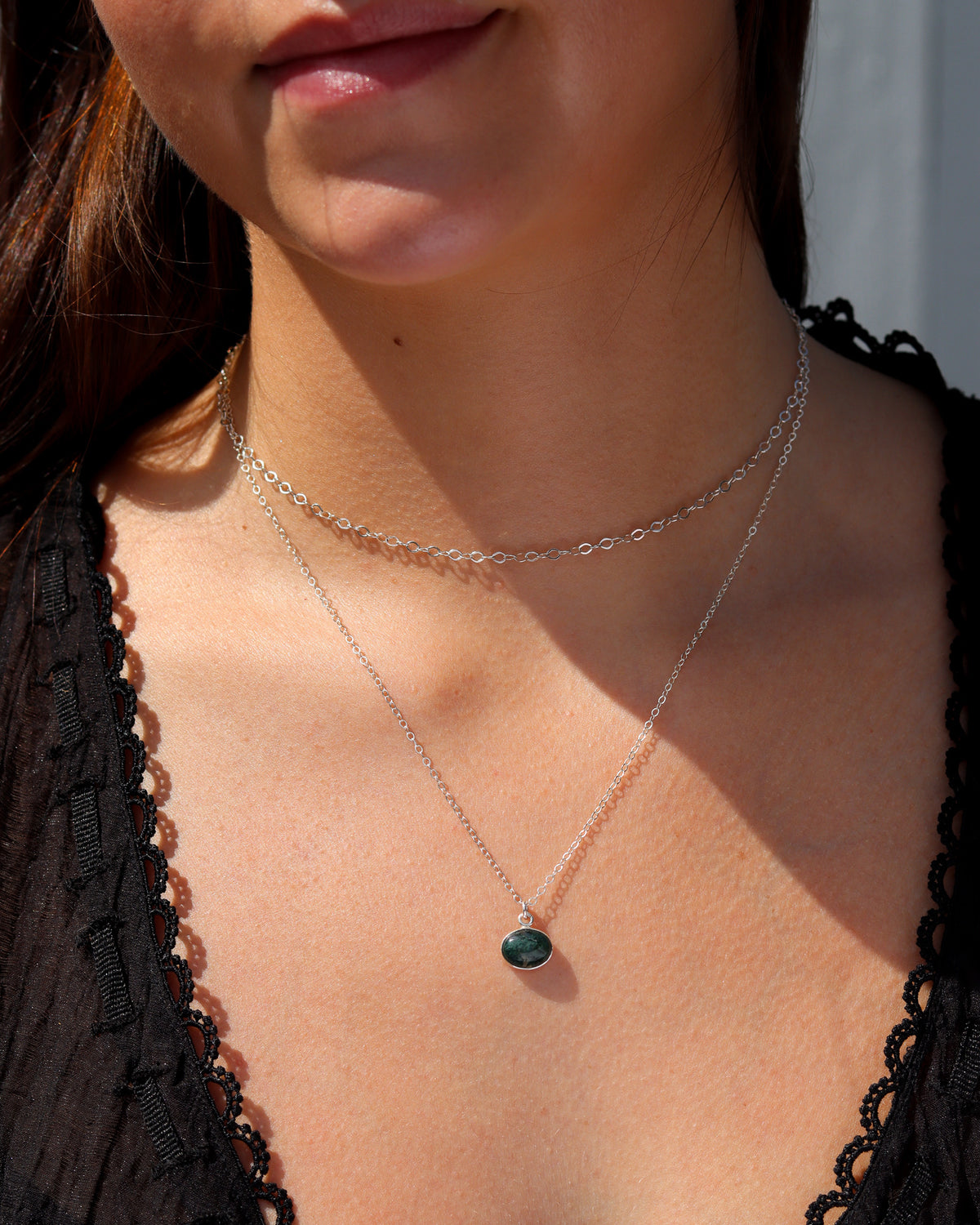 Layered silver necklaces featuring a delicate oval-link chain and a green gemstone pendant, styled in warm sunlight.