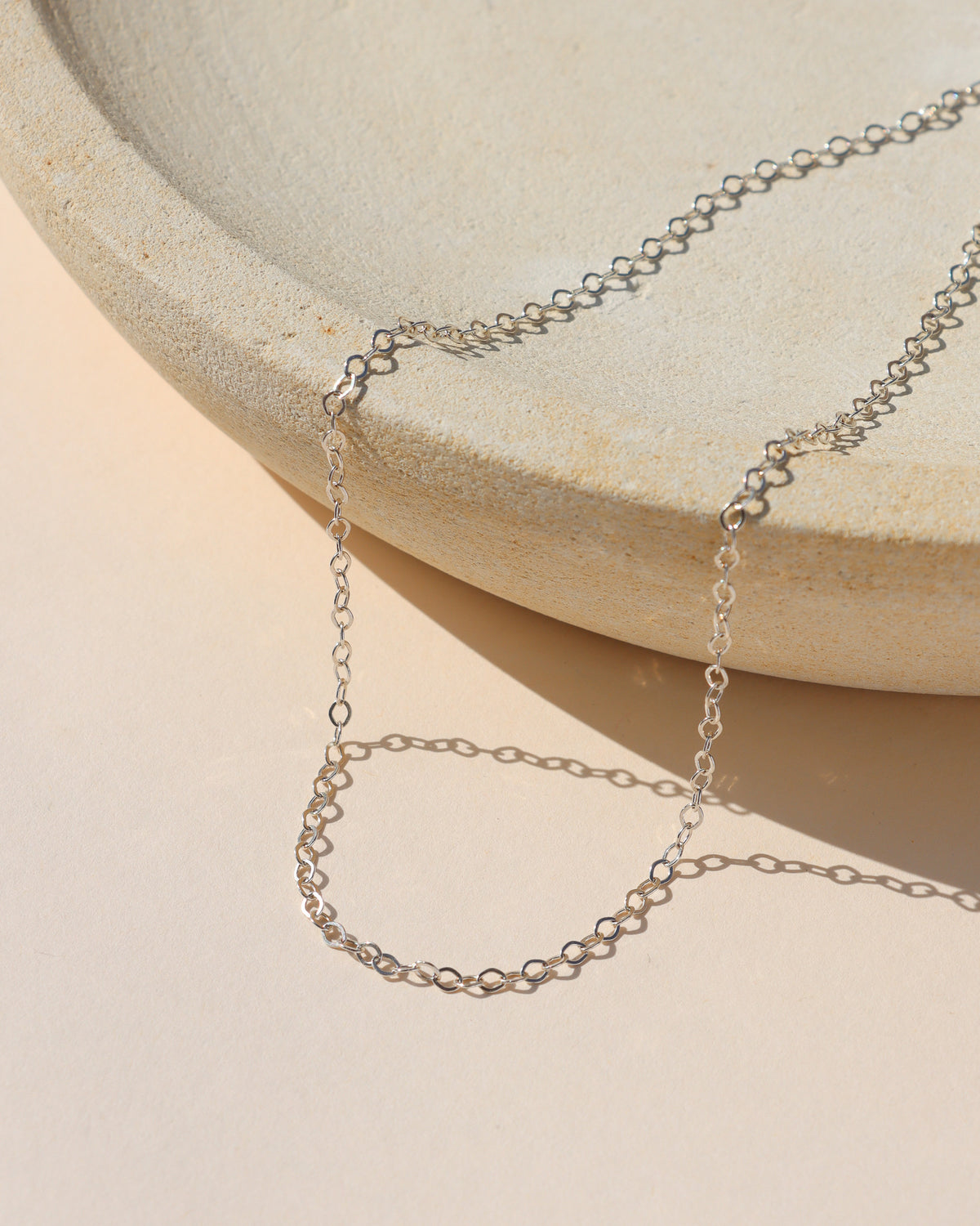 Delicate sterling silver oval-link chain necklace displayed on a neutral surface in warm sunlight.