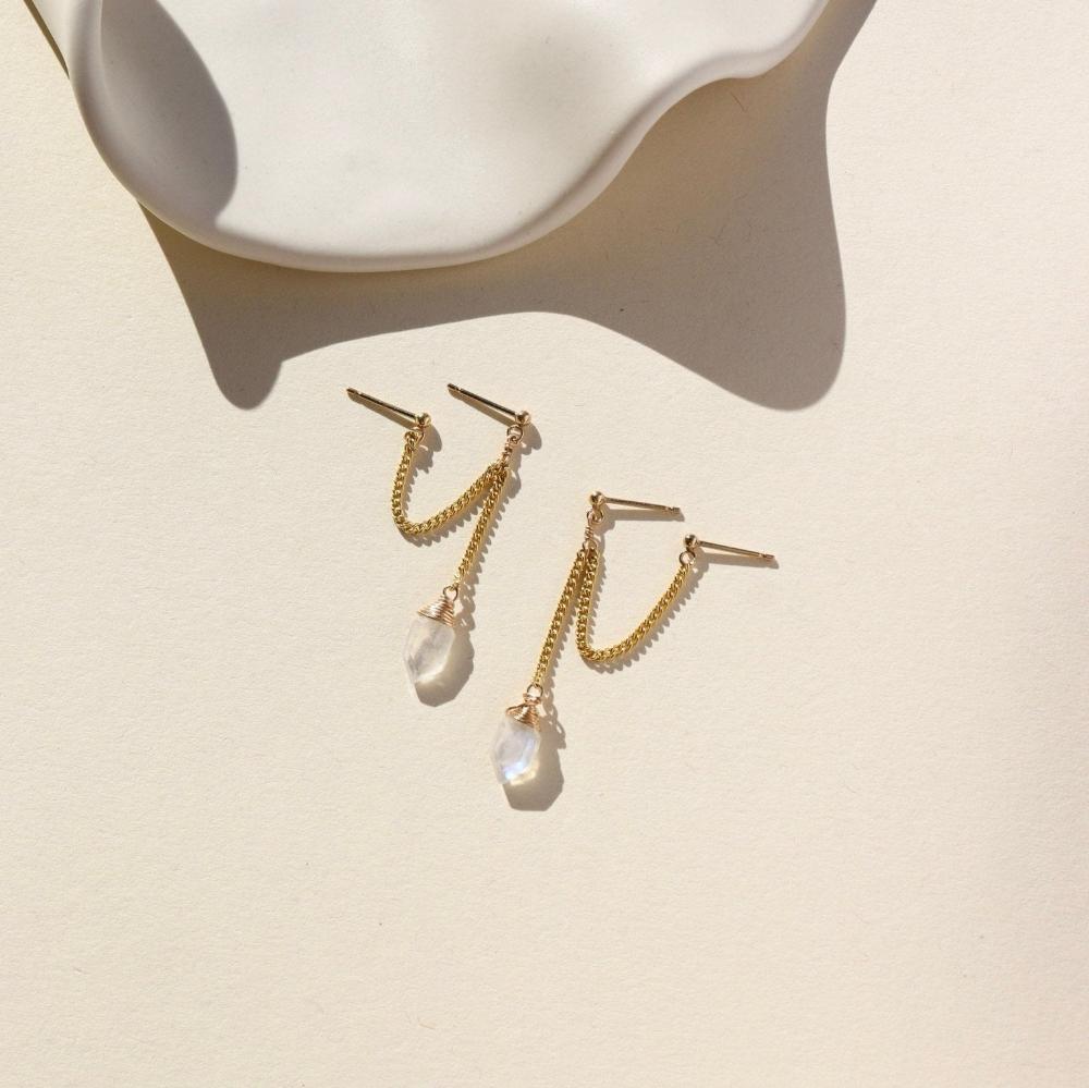 Two small studs with gold curb chain and a hexagon moonstone attached to the end of each earring. Earrings are displayed on a cream backdrop. 