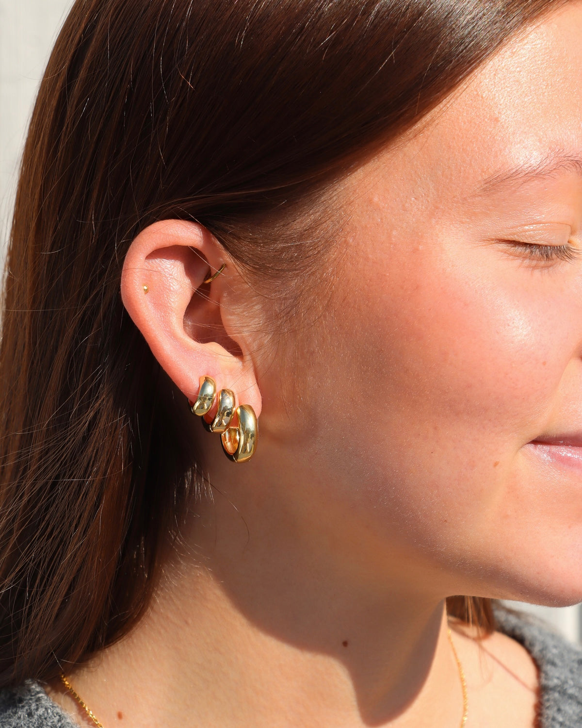 Close-up of a model wearing three polished gold Luxe hoops in Mini, Classic, and Hoop sizes, styled together in multiple lobe piercings with delicate gold cartilage jewelry in warm natural sunlight.