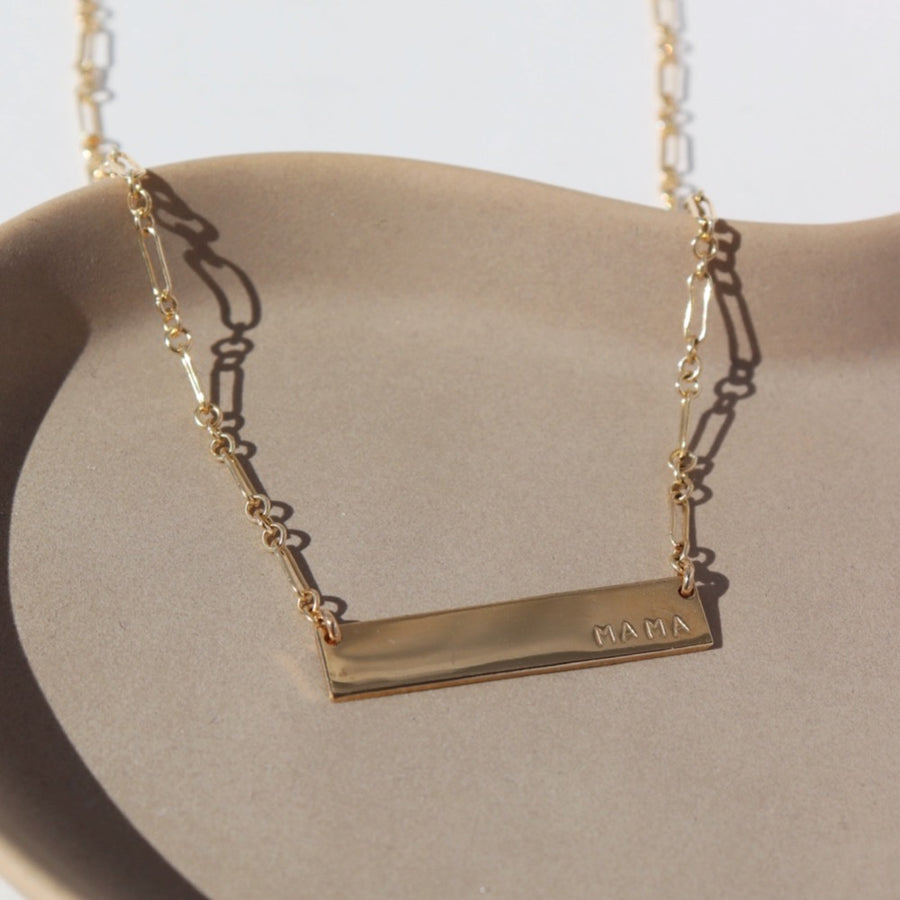 14k gold fill polished bar nameplate necklace with the word "Mama" stamped on the lower left side. The necklace hangs from a gold filled chain with delicate long and short links and is positioned in the sunlight on a ceramic plate.