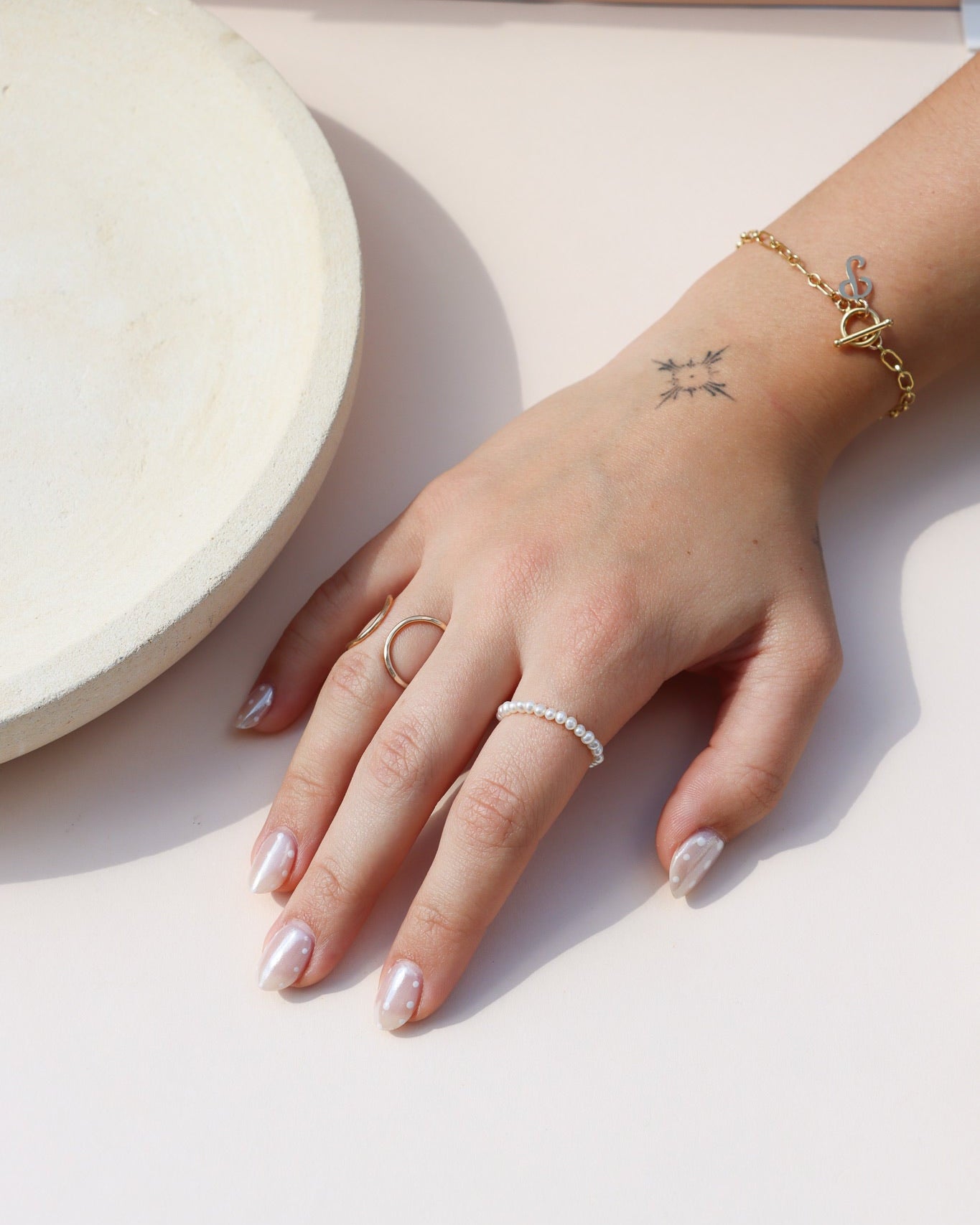 A close-up of a hand modeling a small seed pearl ring, a gold double-circle ring, and a gold chain toggle bracelet with a pendant, resting beside a ceramic dish.