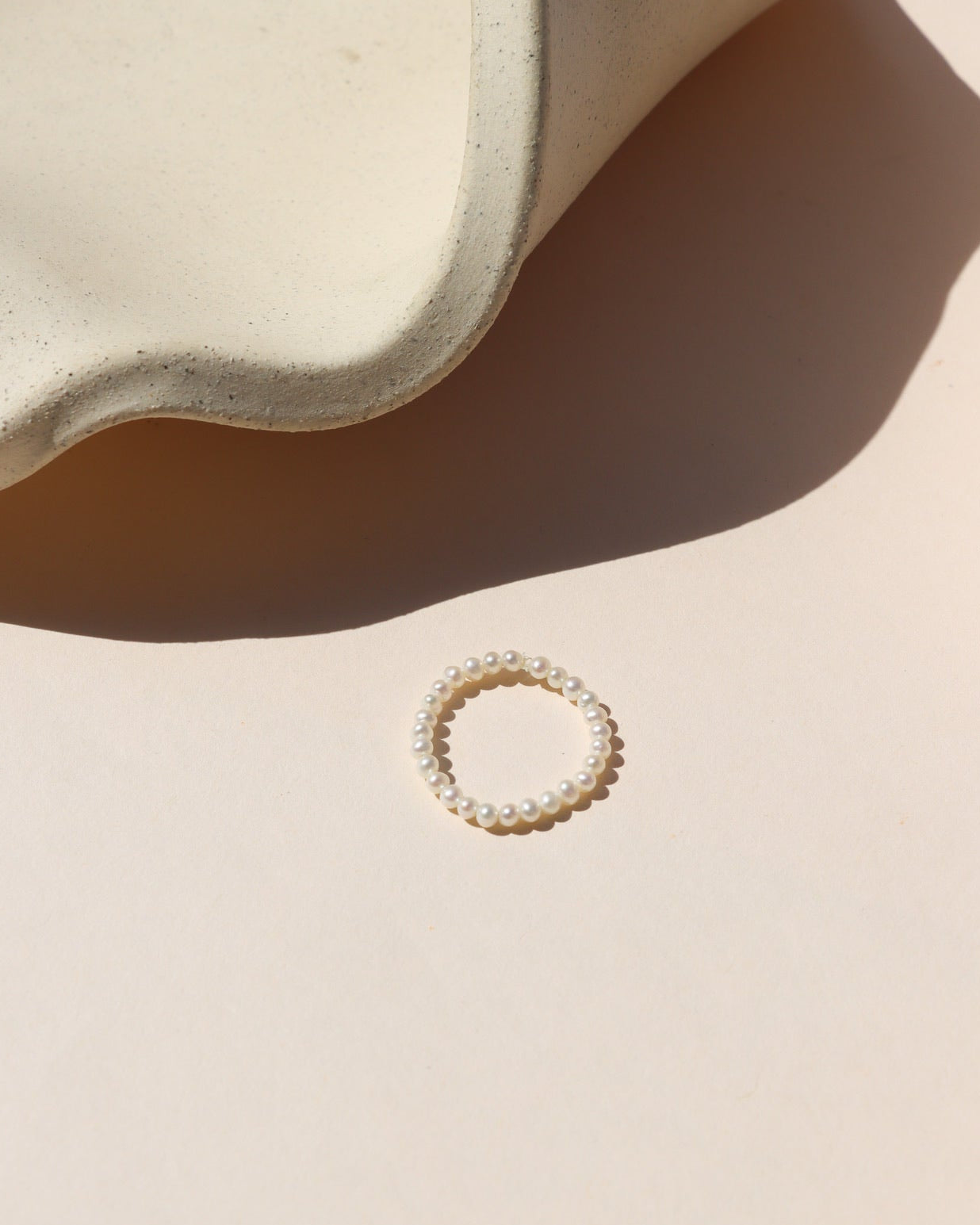 Beaded hand-strung pearl stacking ring laying on a beige surface in the sunlight.