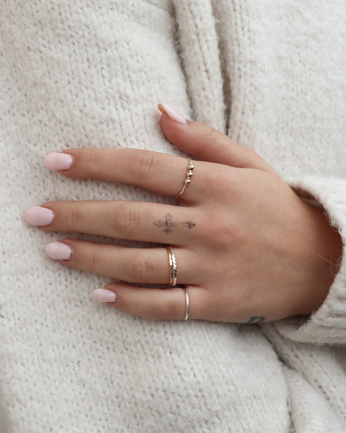 Close-up of a model's hand wearing layered Token Jewelry gold and silver stacking rings, including a delicate three-bead ring, textured bands, and a minimalist silver ring, styled against a soft cream knit sweater.