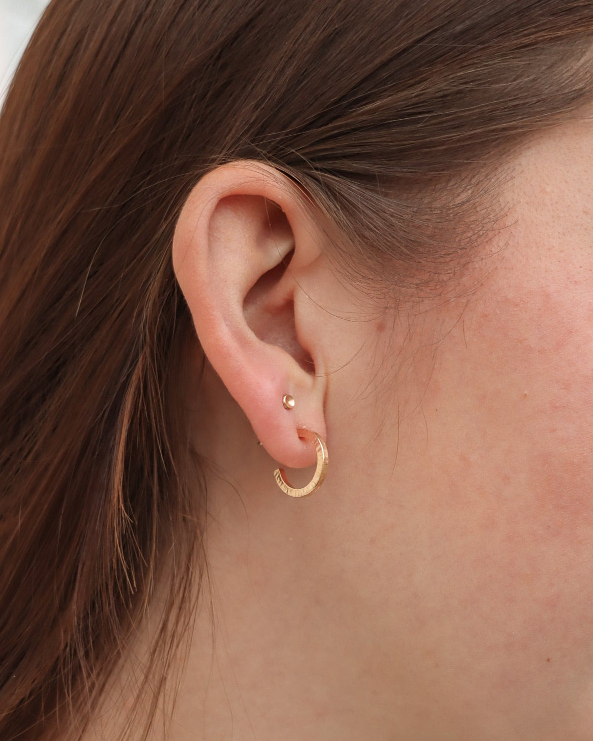 Side view of a model wearing small textured gold hoop earrings paired with a petite gold stud earring.