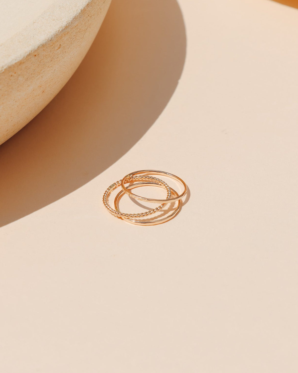 Minimalist stackable gold ring set—featuring a smooth band, a twisted rope band, and a textured band—resting on a warm peach-toned surface with ceramic props and soft sunlight.