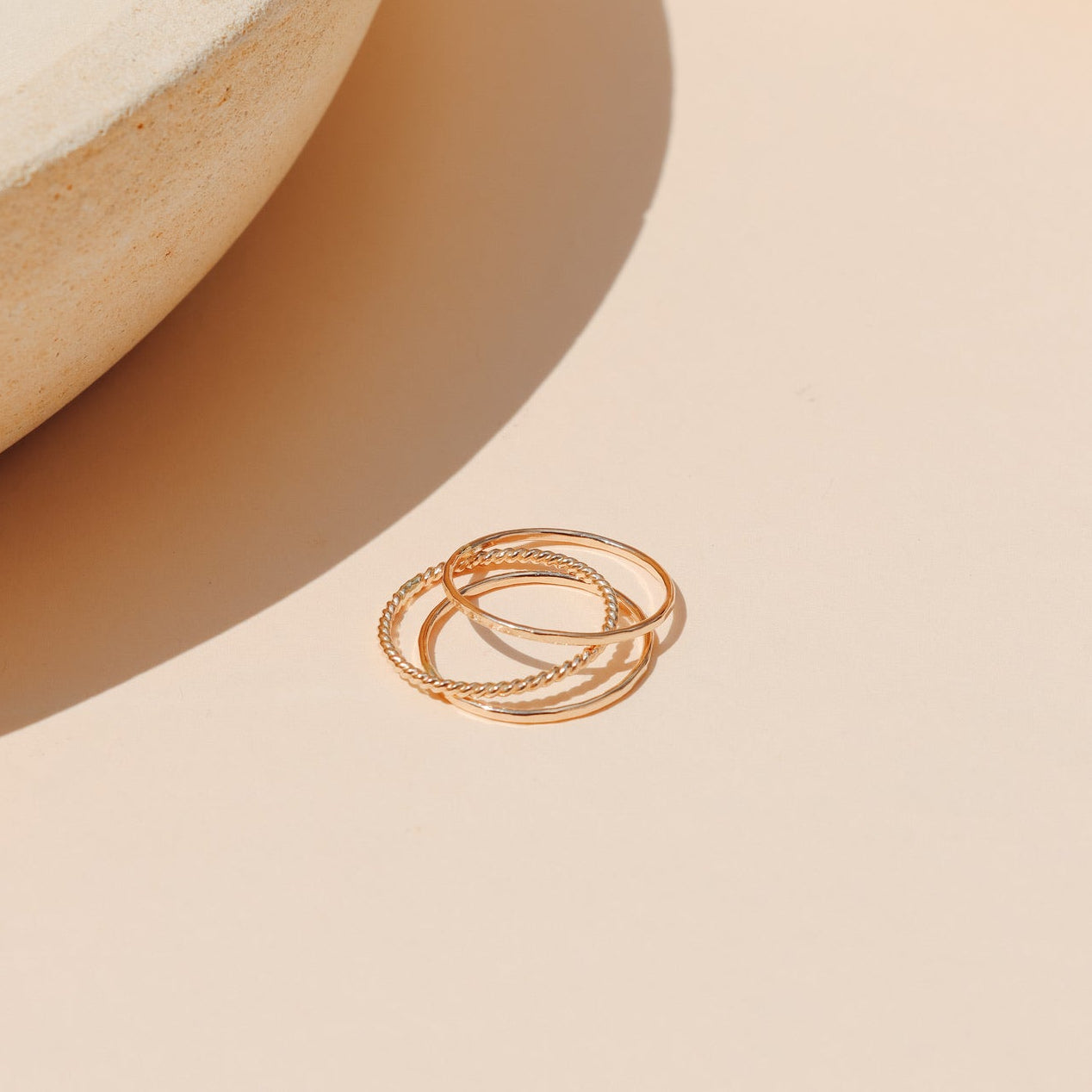 Minimalist stackable gold ring set—featuring a smooth band, a twisted rope band, and a textured band—resting on a warm peach-toned surface with ceramic props and soft sunlight.