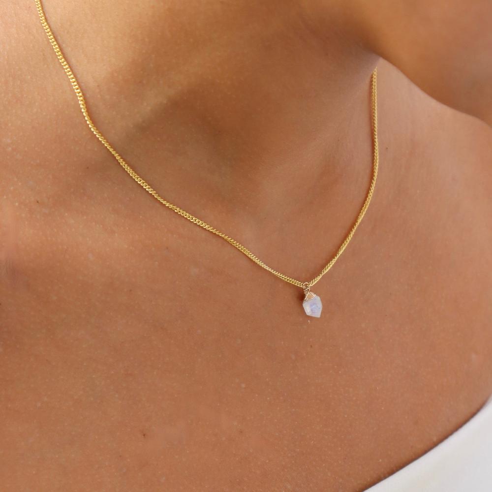 Model wearing the lumi necklace. Necklace features a gold curb chain with a moonstone hanging in the center. Model is wearing a white tube top. 