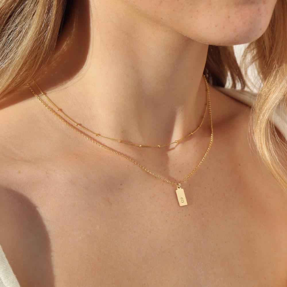 Red head model wearing the mini monogram tag necklace. This necklace features a gold rolo chain with a small stamped &quot;B&quot; rectangle pendant. 