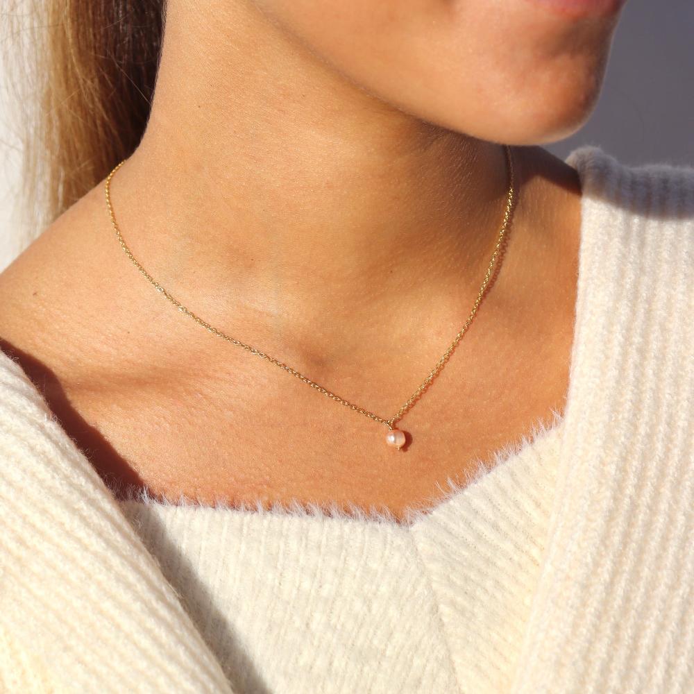 Brunette model wearing the pink pearl necklace. This necklace features a dainty gold chain with a small pink pearl hanging at the end. 