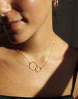 Brunette model wearing the unity necklace. This necklace features a dainty gold chain with a large and small circle connecting in the center. 