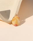 Oval golden yellow and cream stone pendant with a soft red accent, set in gold and displayed beside a textured cream ceramic form.
