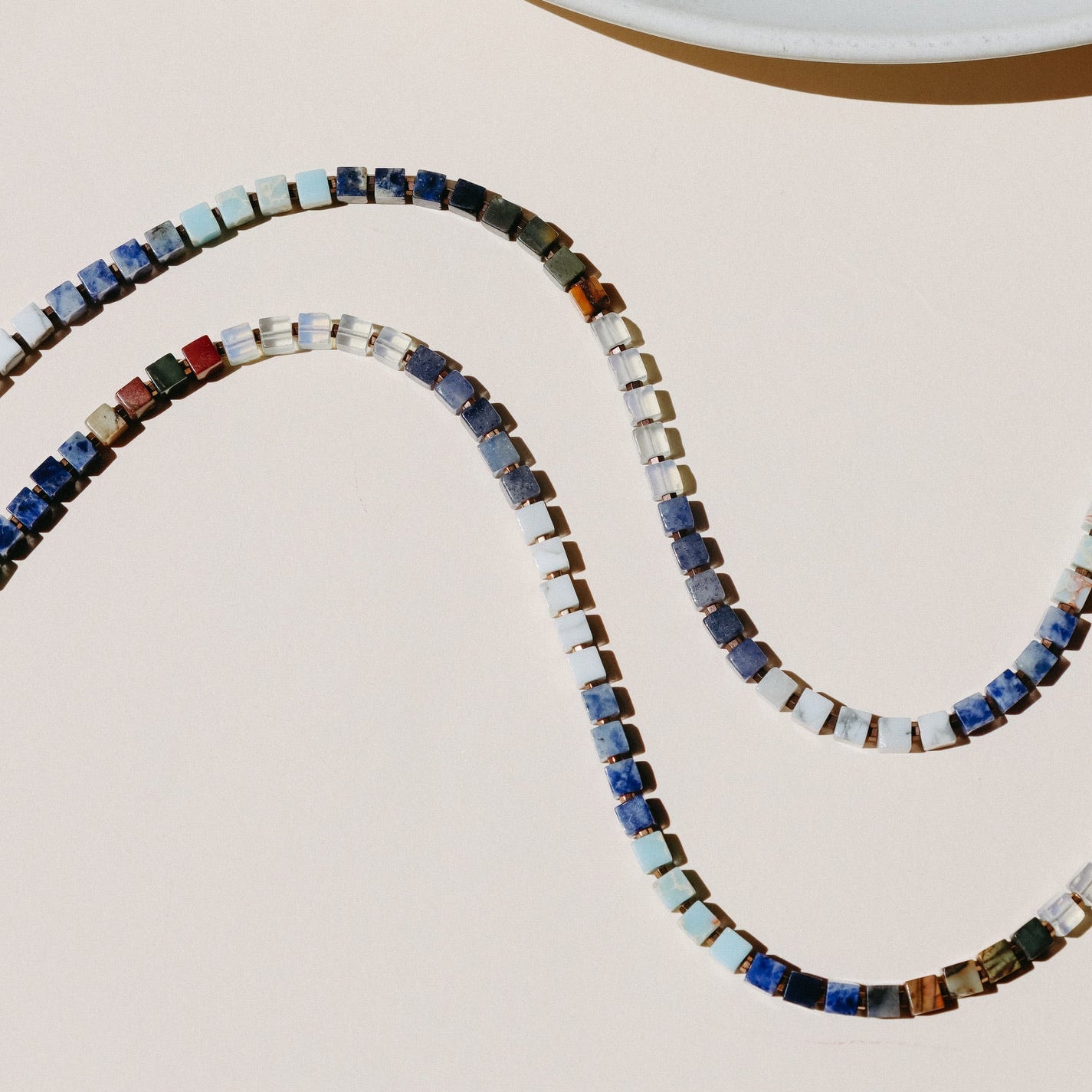 Cubed beaded necklace of various blues and earthtones laid organically on a beige background in the sunlight