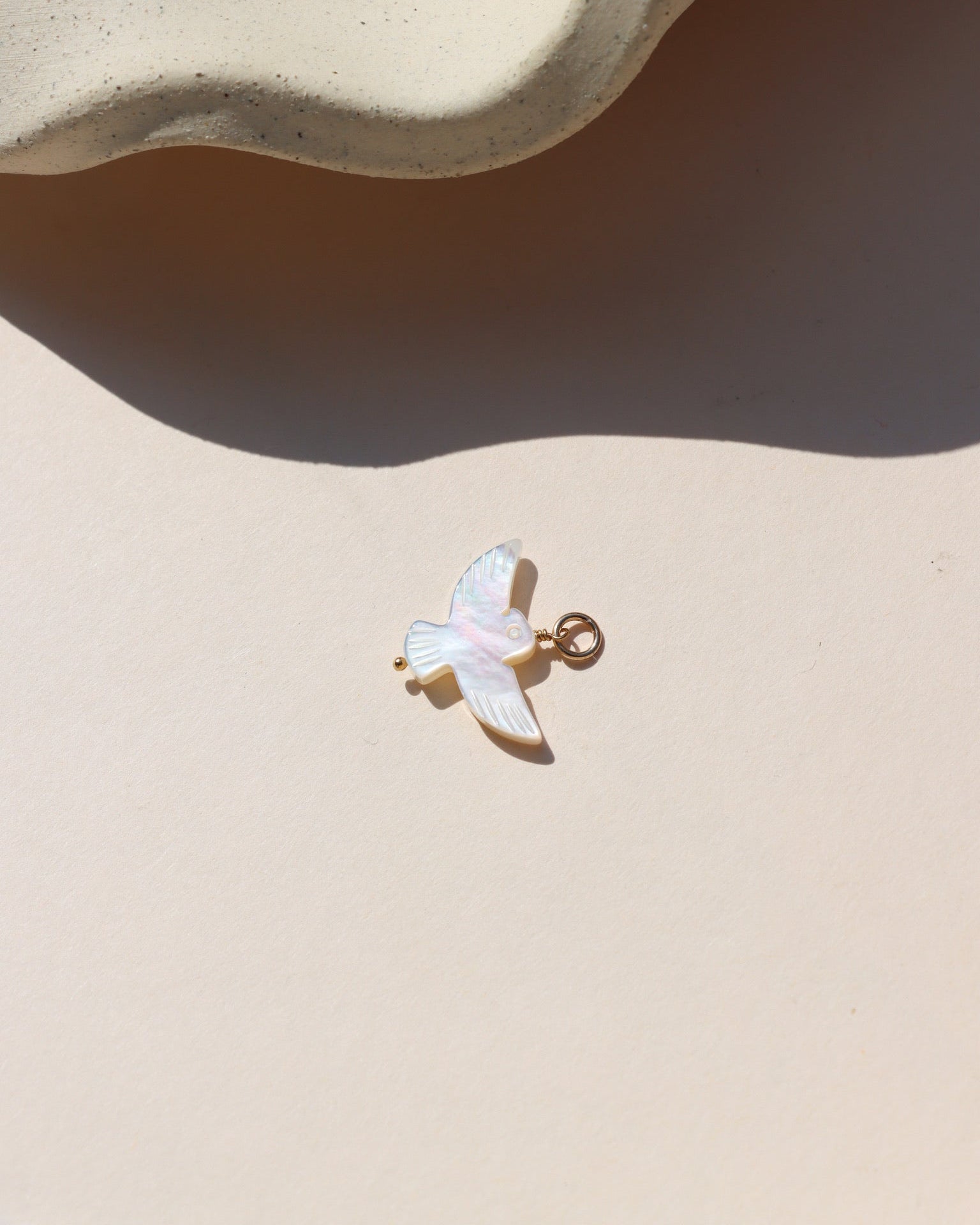 A small iridescent mother-of-pearl bird charm with a gold-toned loop, photographed on a cream-colored surface in sunlight.