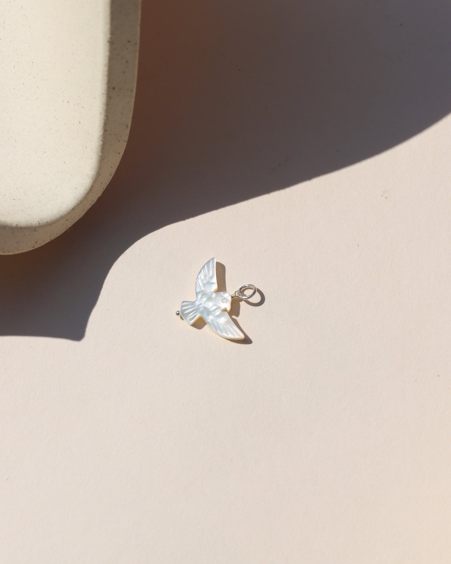 A small iridescent mother-of-pearl bird charm with a sterling silver loop, photographed on a cream-colored surface in sunlight, casting a soft shadow.