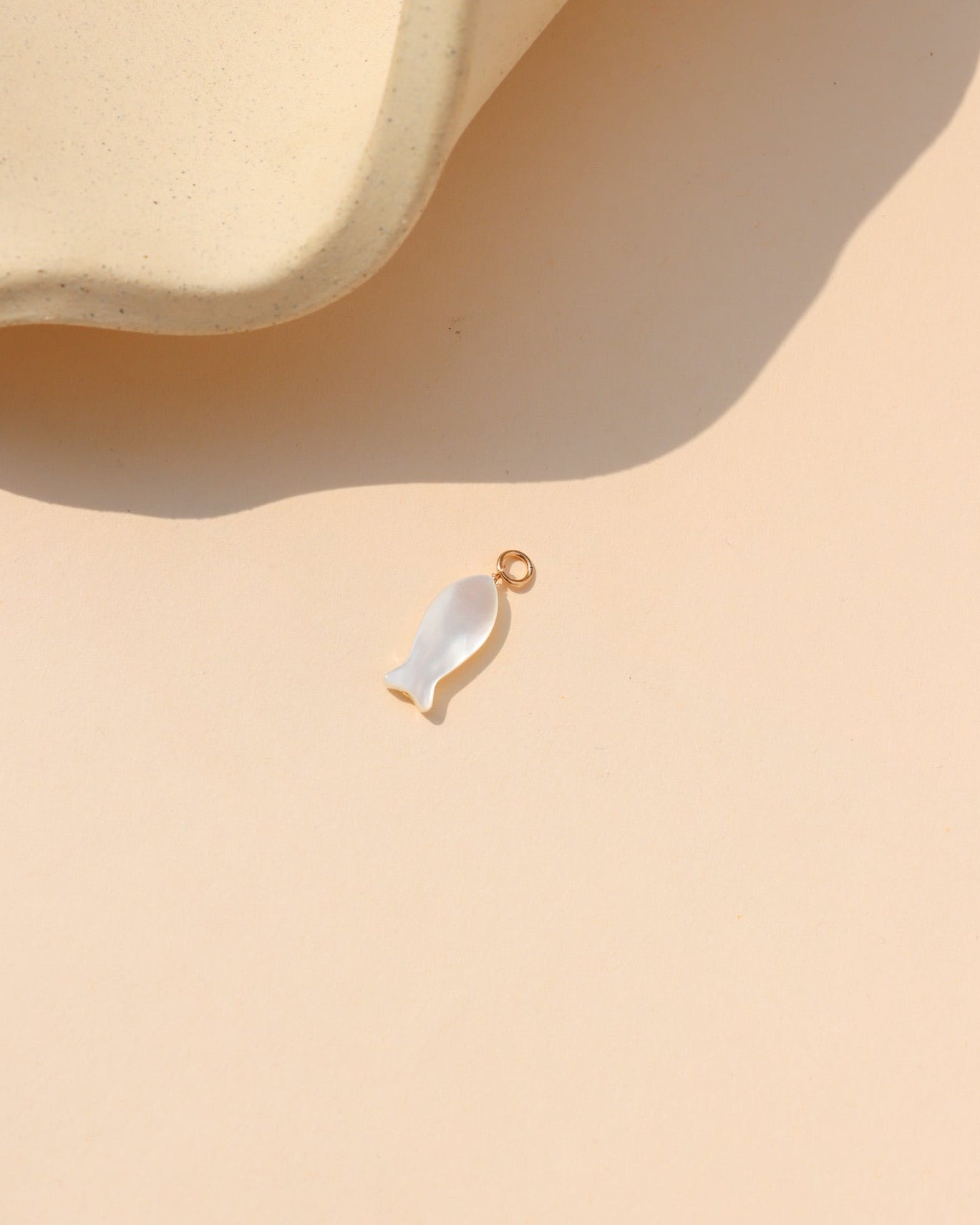  Mother-of-pearl fish charm with gold loop, photographed on a warm cream background beside a ceramic form.