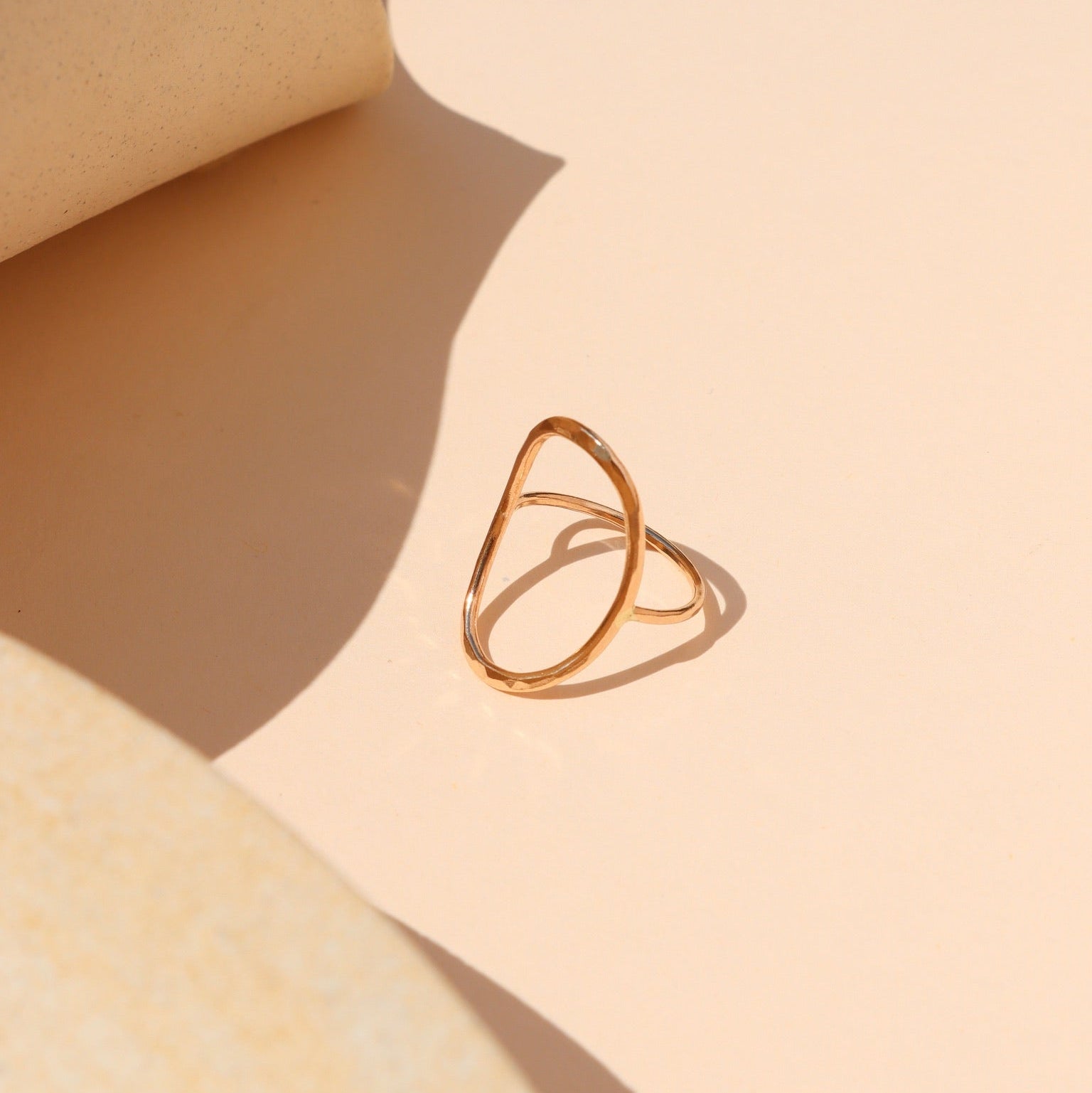 Minimalist gold ring with an open oval outline design displayed on a peach-beige surface, surrounded by curved neutral props and dramatic warm sunlight shadows.