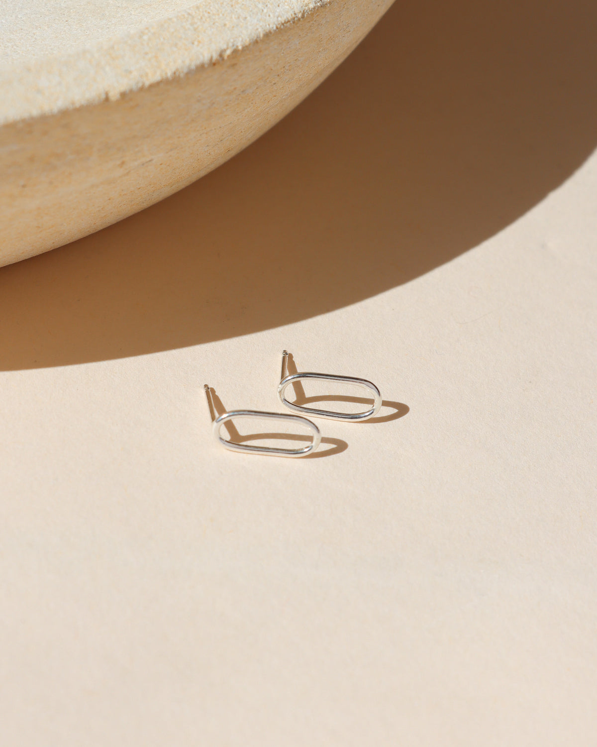 Sterling silver open oval stud earrings displayed on a neutral surface in warm sunlight.