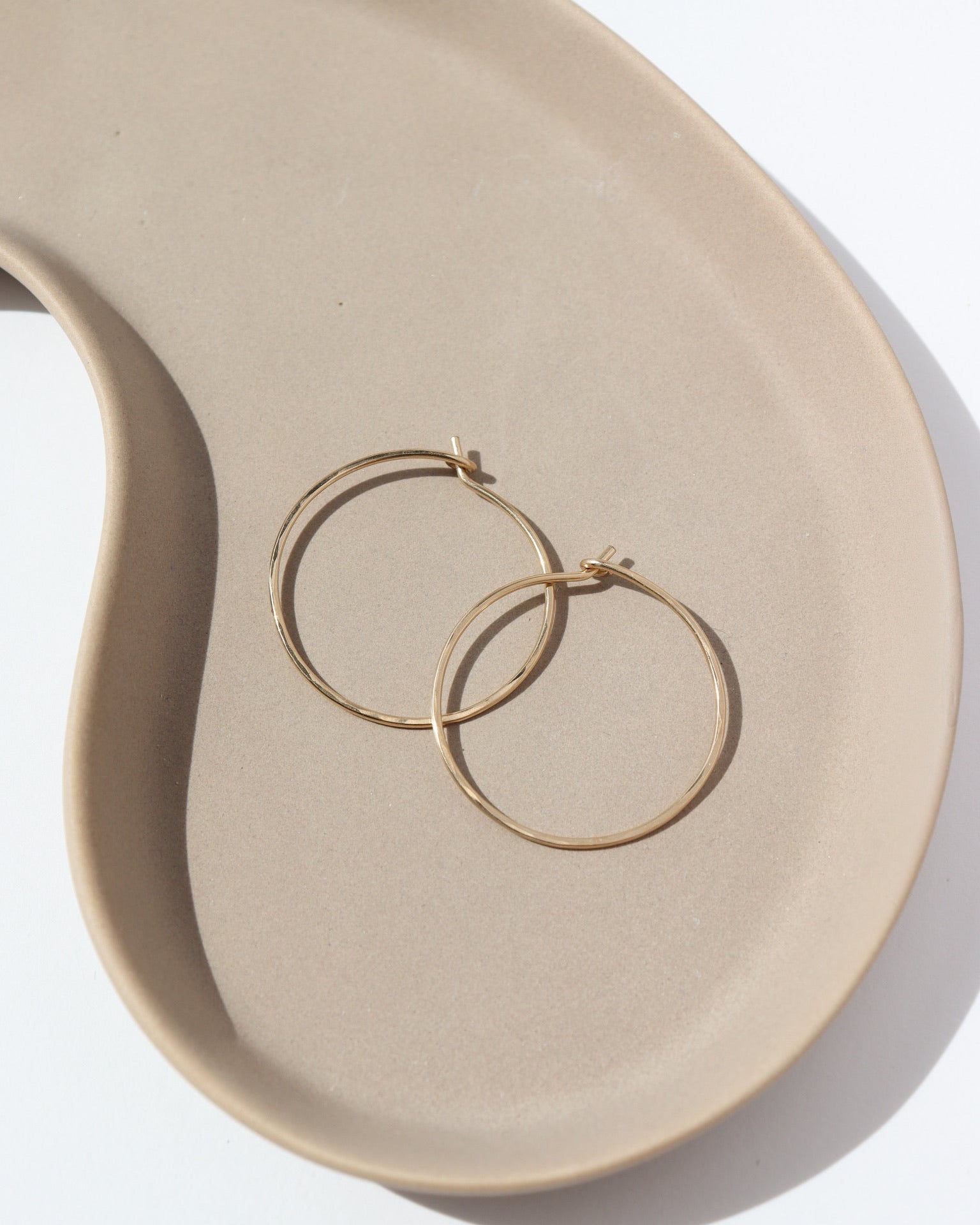 14k solid gold Organic Hoops by Token Jewelry. The hoops are a circle shape with hammered solid gold wire, photographed on a tan ceramic dish