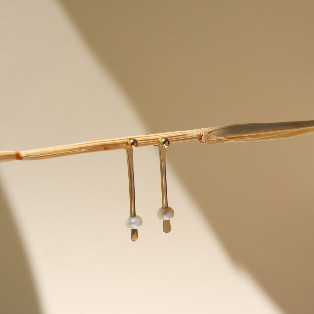 A 14k gold fill drop down post with a pearl on each ear jacket, the earrings are displayed on a cream back drop in the sunlight. 