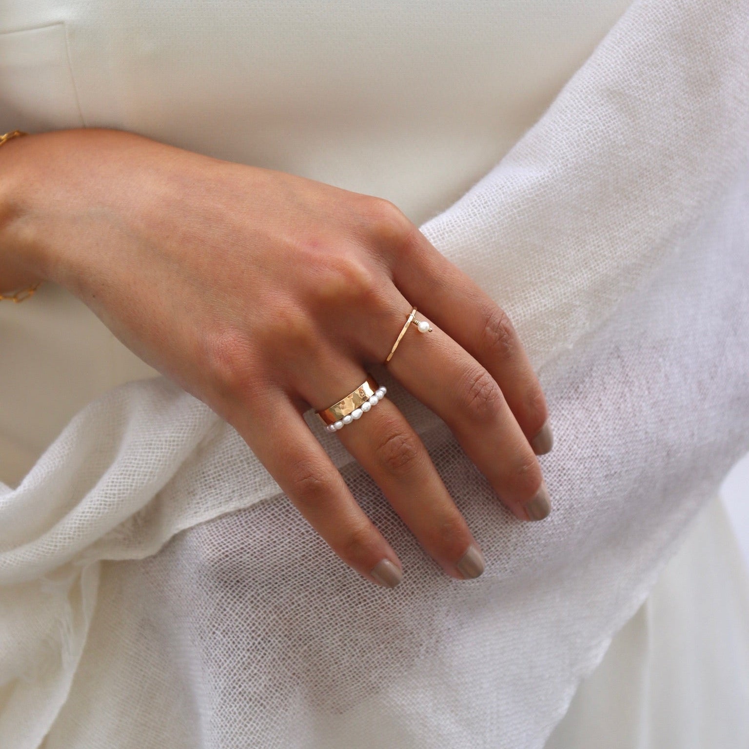 Close-up of a model wearing elegant gold and pearl rings against a white outfit, featuring a wide hammered gold band stacked with a mini pearl bead ring on her ring finger, and a thin gold band with a tiny dangling pearl drop on her middle finger.