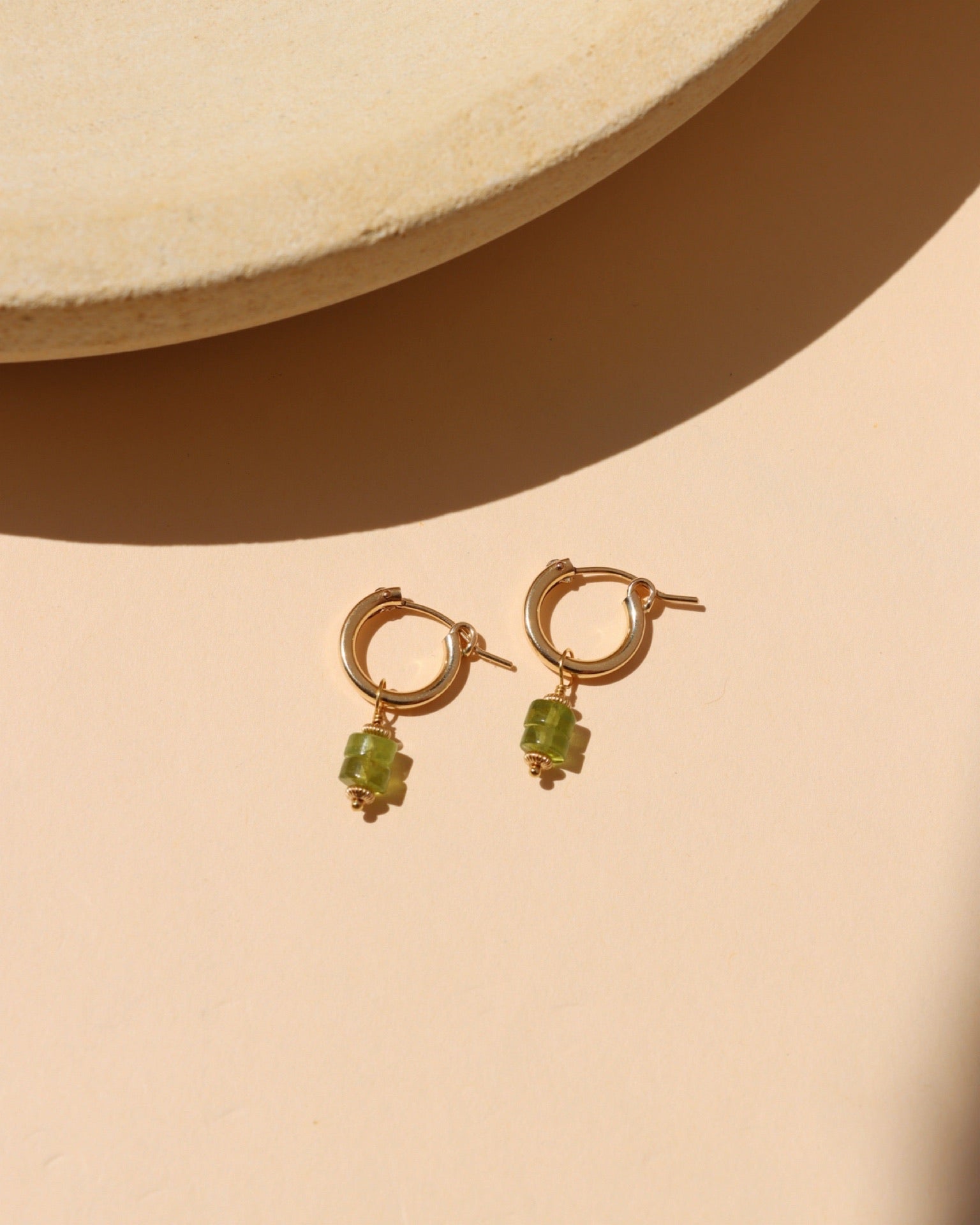 Petite gold hoop earrings with natural green peridot gemstone drops, photographed in warm sunlight.