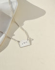 Sterling Silver necklace with a thin rolo chain with a silver smooth rectangle pendant with a "J" and a "T" stamped in the middle of it. The necklace is displayed on a cream backdrop. 