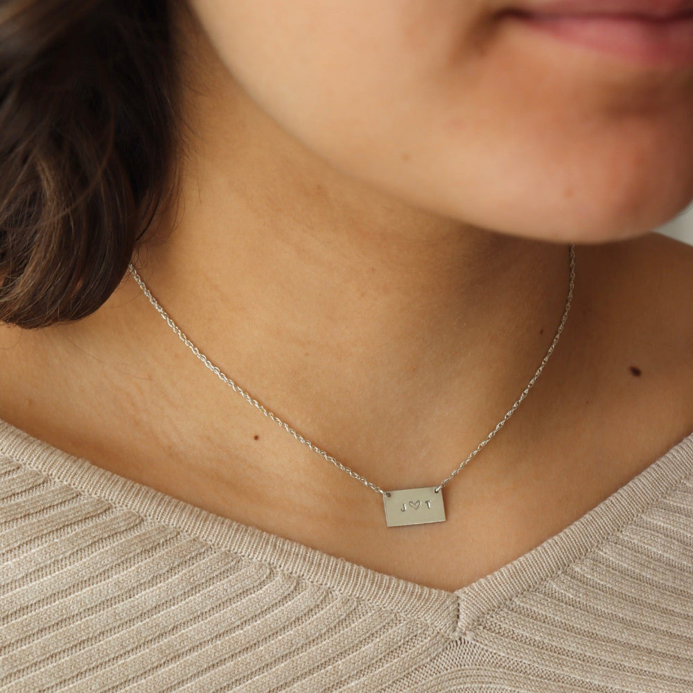 Model wearing sterling Silver necklace with a thin rolo chain with a silver smooth rectangle pendant with a "J" and a "T" stamped in the middle of it.