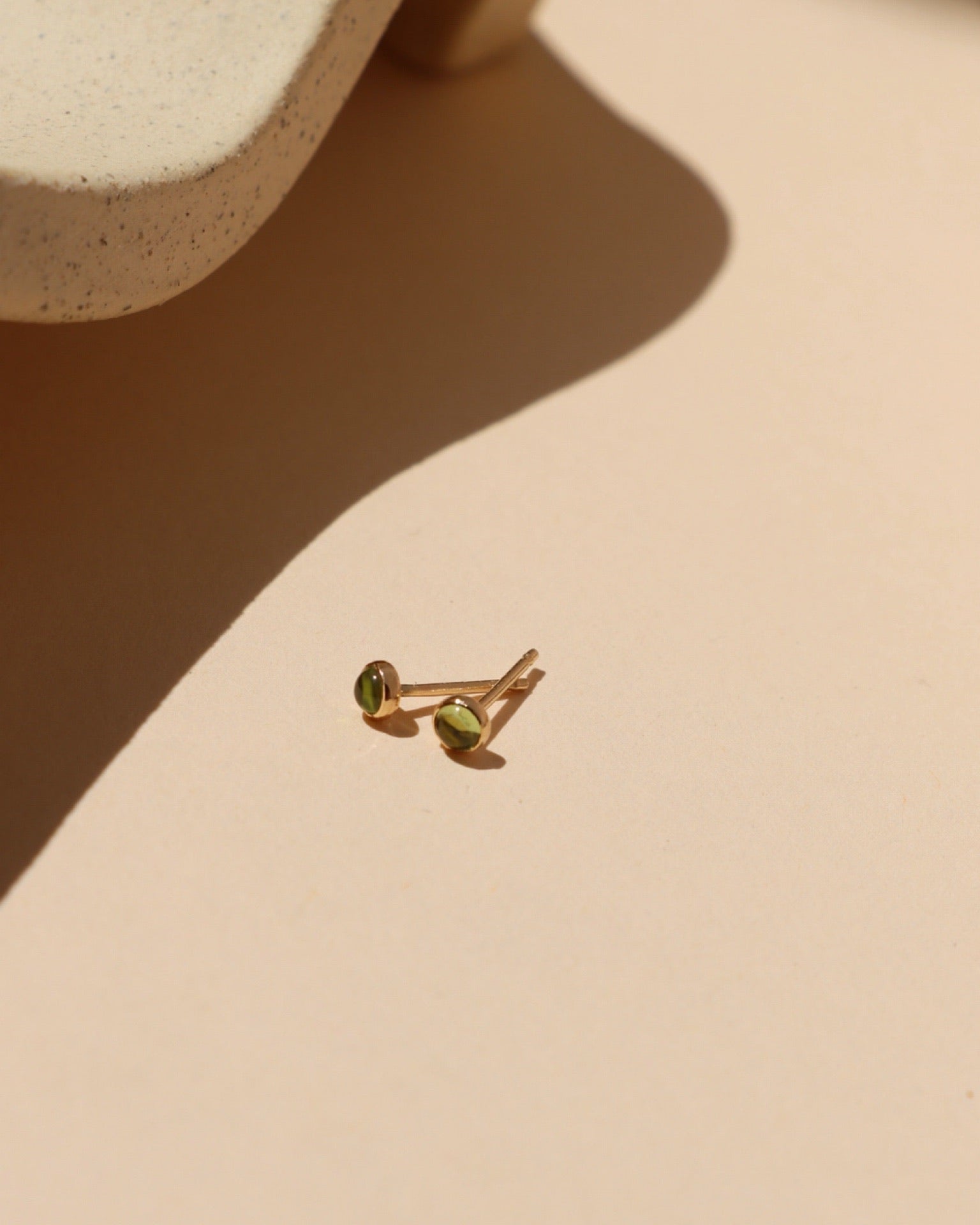 Petite round green peridot gemstone stud earrings in gold settings, August birthstone jewelry photographed in warm sunlight.