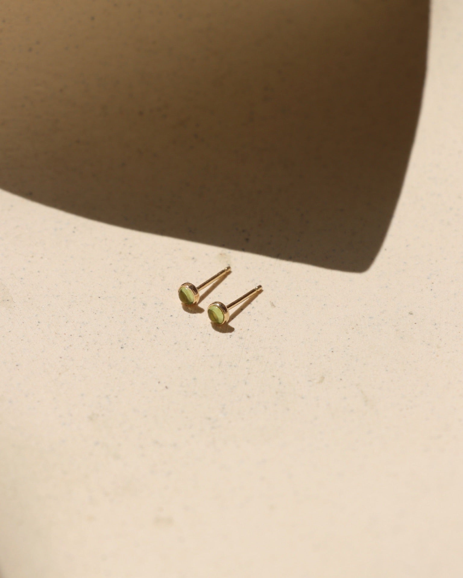 Petite round green peridot gemstone stud earrings in gold settings, August birthstone jewelry photographed in warm sunlight.