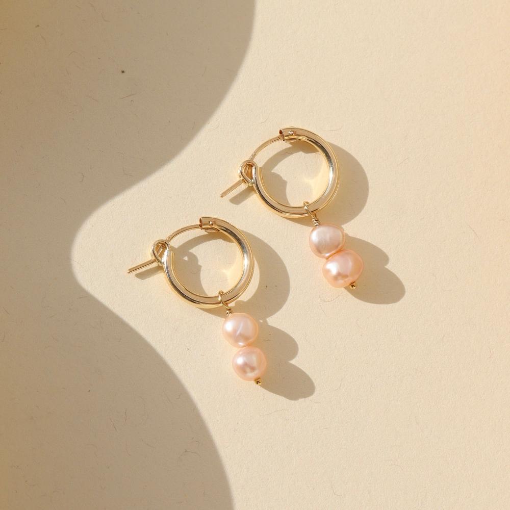 Small gold hoops featuring two pink pearls dangling from them. Earrings are displayed on a cream backdrop.