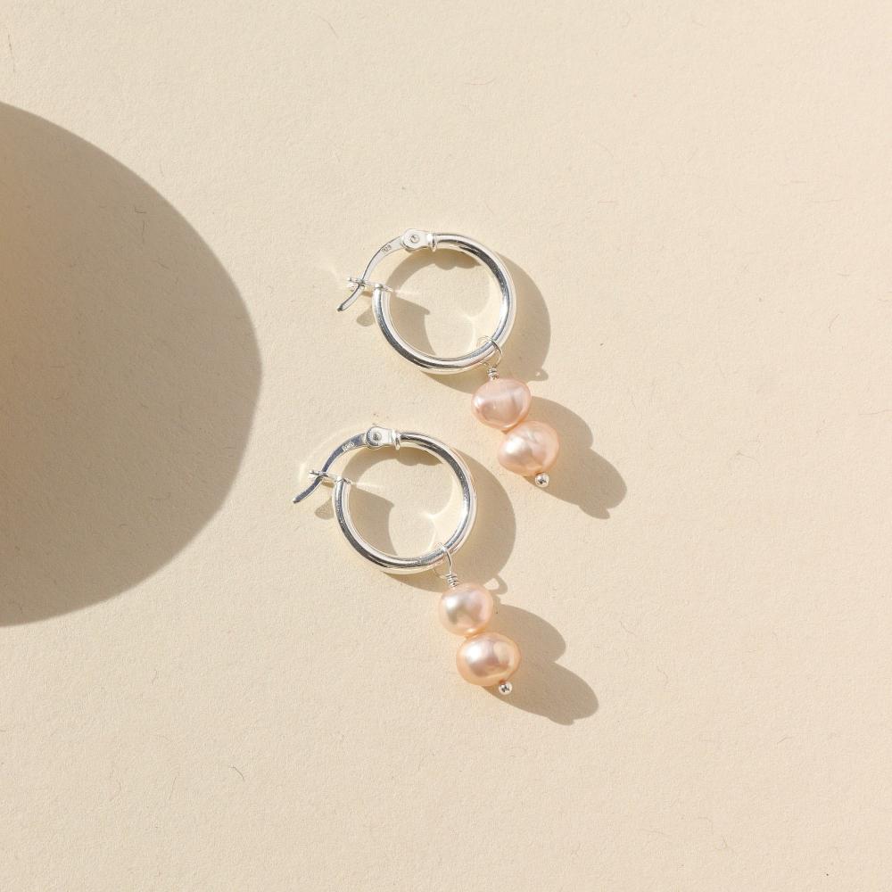 Small silver hoops featuring two pink pearls dangling from them. Earrings are displayed on a cream backdrop.