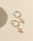 Small silver hoops featuring two pink pearls dangling from them. Earrings are displayed on a cream backdrop.