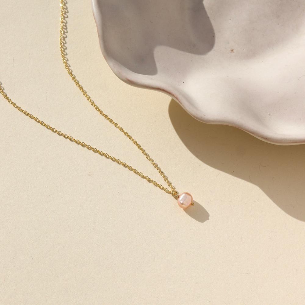 Delicate gold chain with single pink pearl. Displayed on cream backdrop. 