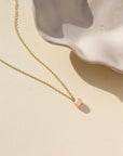 Delicate gold chain with single pink pearl. Displayed on cream backdrop. 