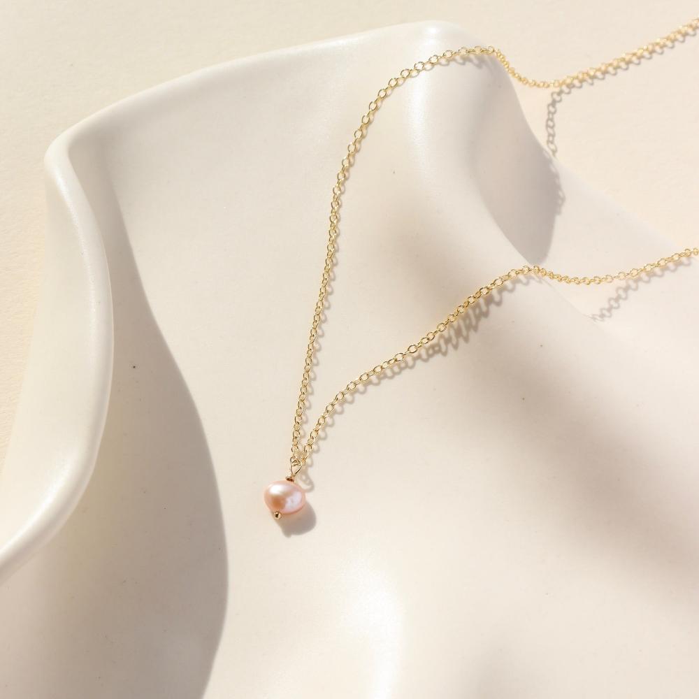 Delicate gold chain with a single pink pearl. Displayed on white dish. 