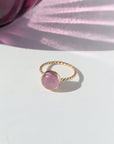 14k gold filled Pippa Ring laid on a white paper in the sunlight. This ring features a sprial band with a pink kunzite gemstone.