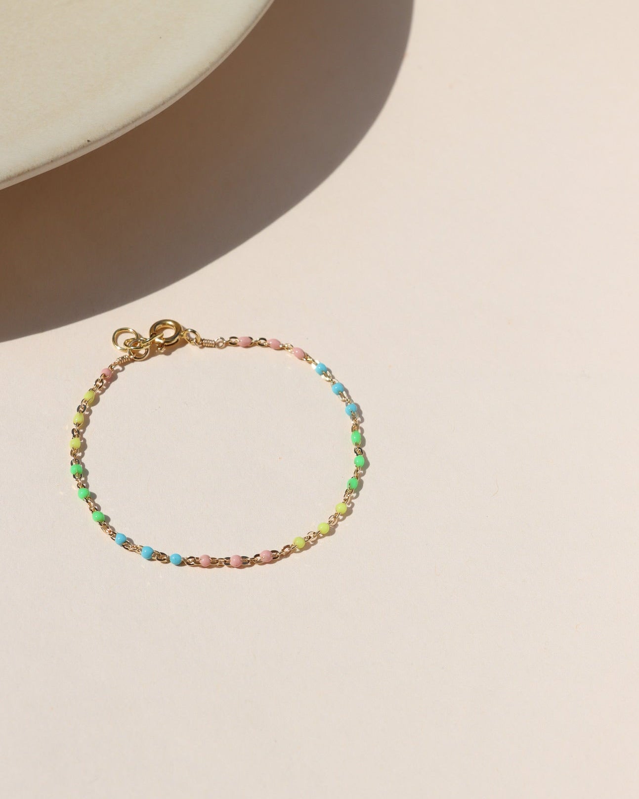 A delicate gold chain bracelet dotted with small rainbow enamel beads, arranged in a circle on a neutral beige background.