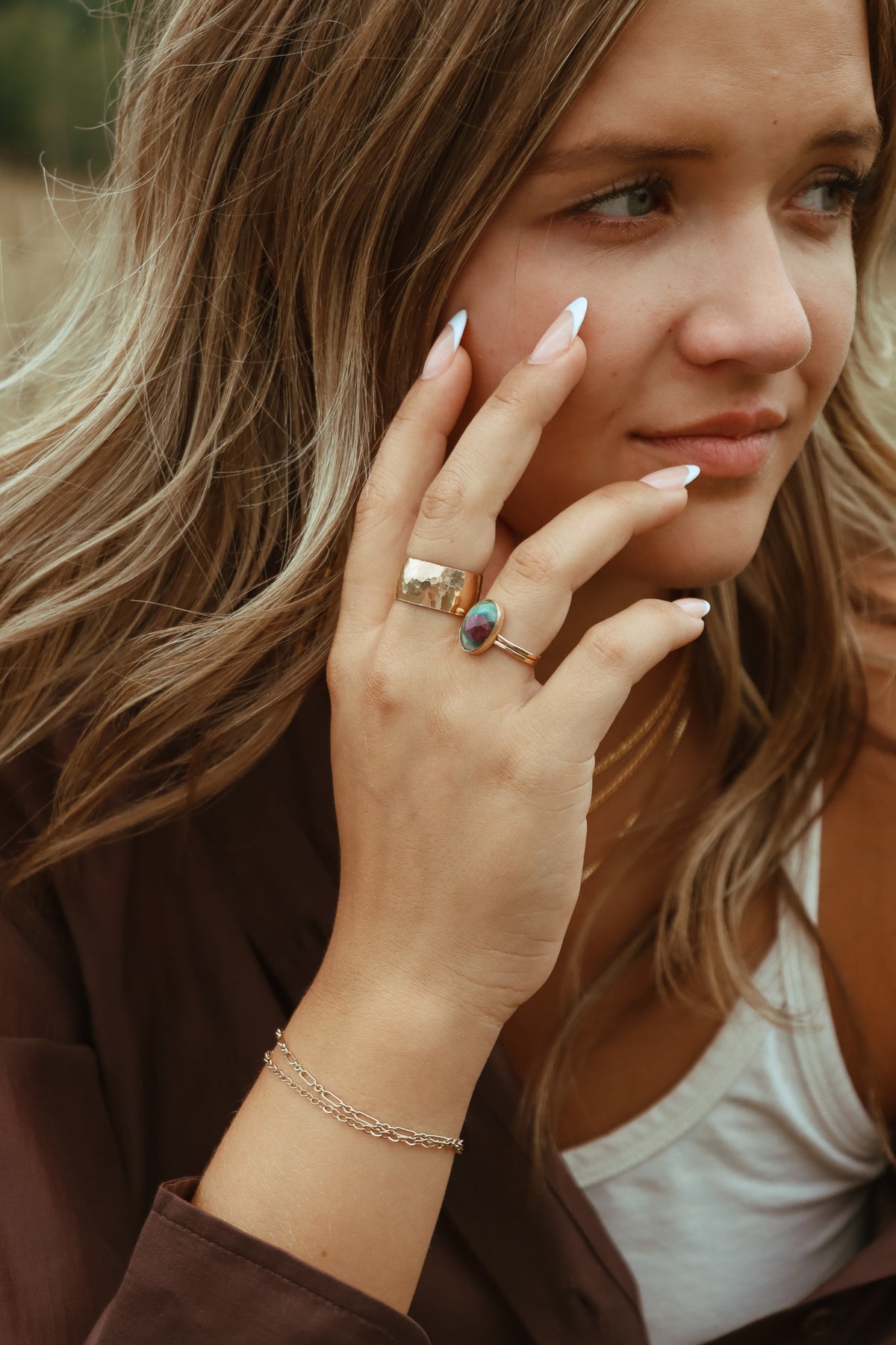 Close-up of a model wearing an oval green and pink gemstone ring, paired with a hammered gold band and layered gold bracelets.