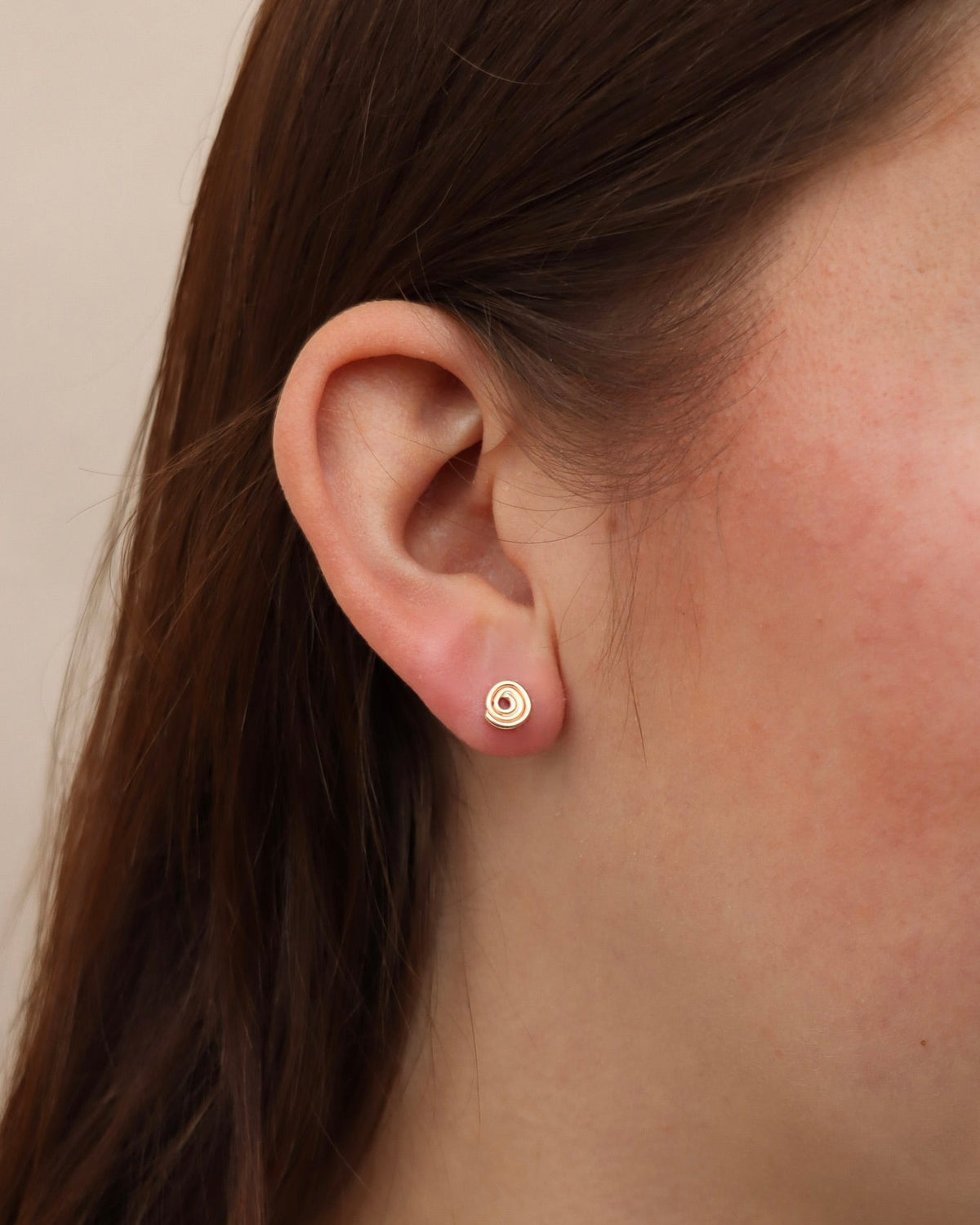 Close-up of a model wearing a small gold spiral stud earring with concentric circular detailing, styled in a single lobe piercing.