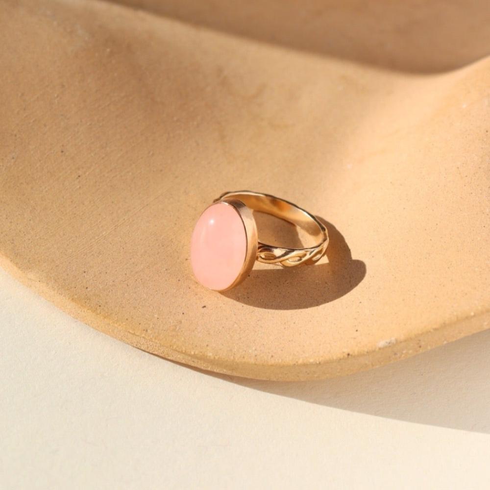 This photo features our rose quartz entwined ring in 14k gold fill. This ring has a 14x10mm  rose quartz gemstone with a thick band with a braided and entwined texture. Ring is displayed on a tan plate. 