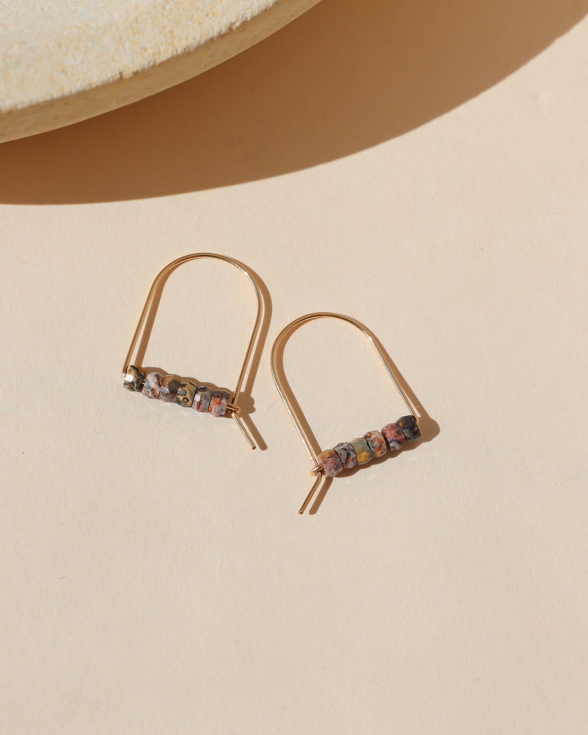 Gold arch earrings with a row of small multicolored stone beads in earthy red, cream, gray, and golden tones, photographed in warm sunlight.