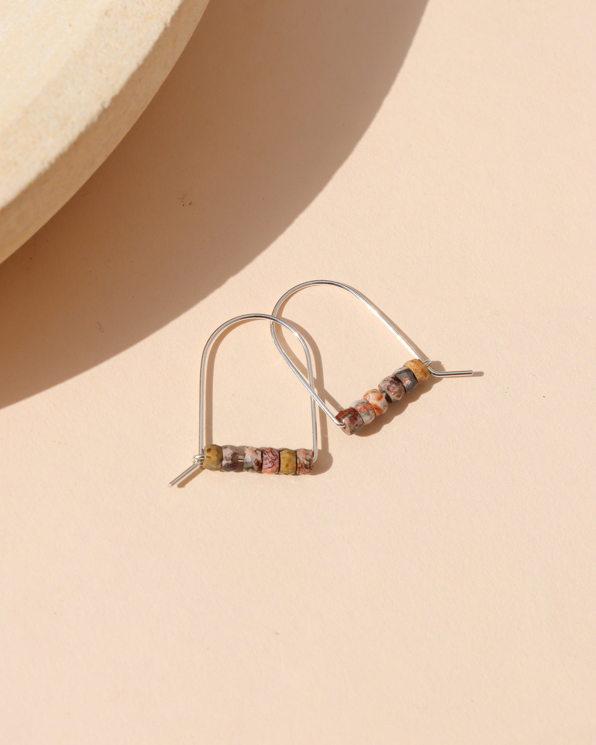 Silver arch earrings with a row of small multicolored stone beads in cream, red, gray, and golden tones, arranged on a cream background in warm sunlight.
