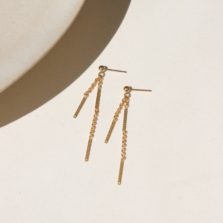 Two 14k gold fill chain stud earrings hanging off a small ball stud, the chain features a bar + link style. The earrings are displayed on a cream back drop in the sunlight.