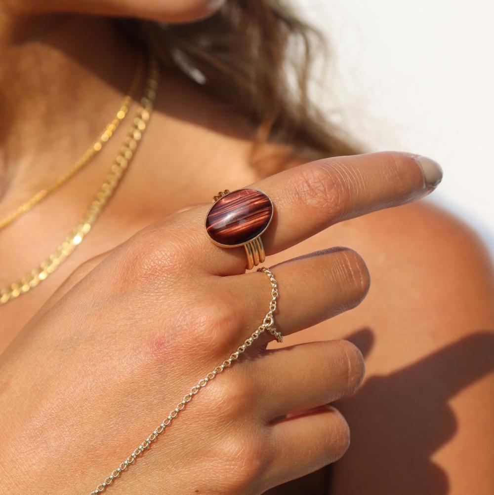The Scarlett Ring, available in 14k gold fill or sterling silver, features a stunning Red Tiger Eye gemstone on a triple 14k gold fill band. It reflect light beautifully as the model wears it along with a hand chain.